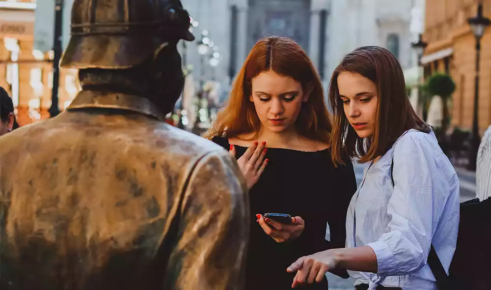 two girls are playing a scavenger hunt on their mobile phones in Budapest, in front of the pre-war policeman statue