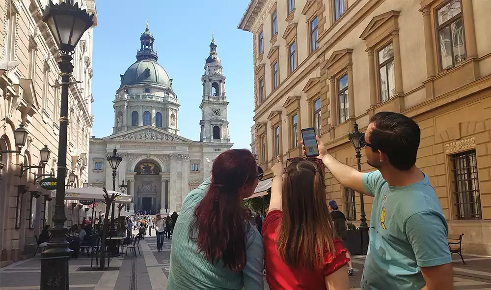 three young people in Budapest are playing a city adventure game while pointing towards St. Stephen's Basilica