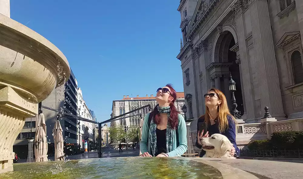 two girls with a dog are sitting on the square in front of Budapest's St. Stephen's Basilica and gazing the fountain