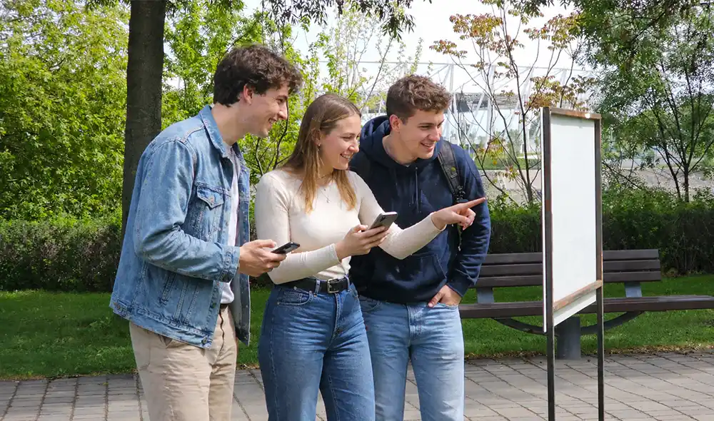 a group of young people at BudaPart standing by a poster, playing the Landventure outdoor escape game on smartphones