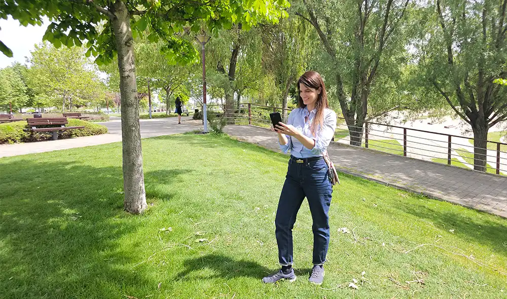 a young woman standing at BudaPart, holding a smartphone and playing the Landventure detective game