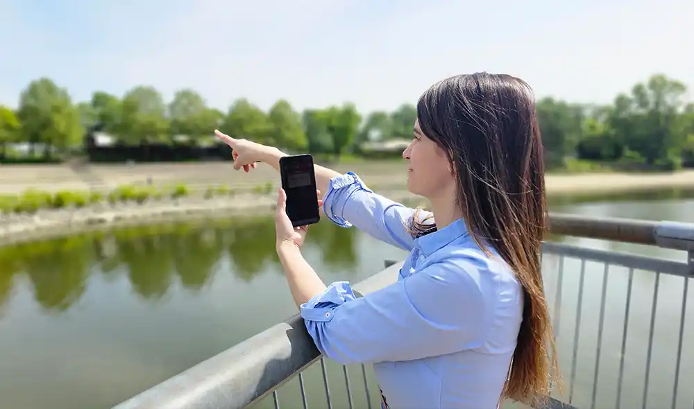 a young woman at BudaPart playing the Landventure city adventure game on her smartphone and pointing into the distance