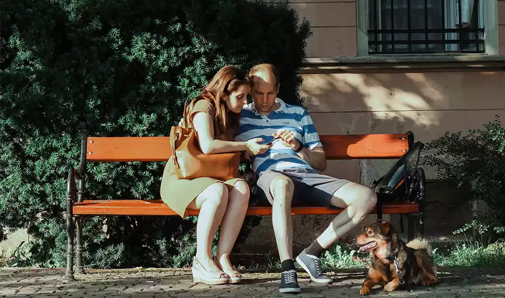 young couple with a dog is playing scavenger hunt on mobile phone while resting on a bench in the Buda Castle District