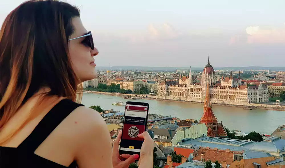 a girl is playing a Landventure scavenger hunt on her mobile phone with the Danube and the Parliament in the background 