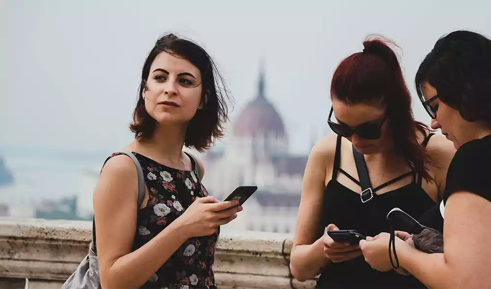 three girls are playing scavenger hunt with mobile phones in front of Buda Castle, with the Parliament in the background