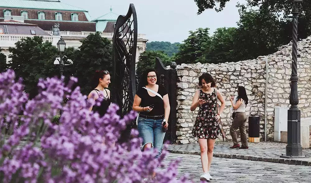 three women in summer outfit are walking with mobile phones in the Buda Castle District