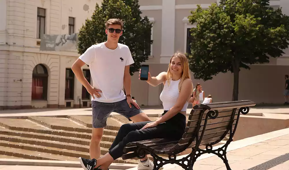 a young couple resting on a bench in downtown Pécs, with the girl showing a smartphone displaying an urban detective game